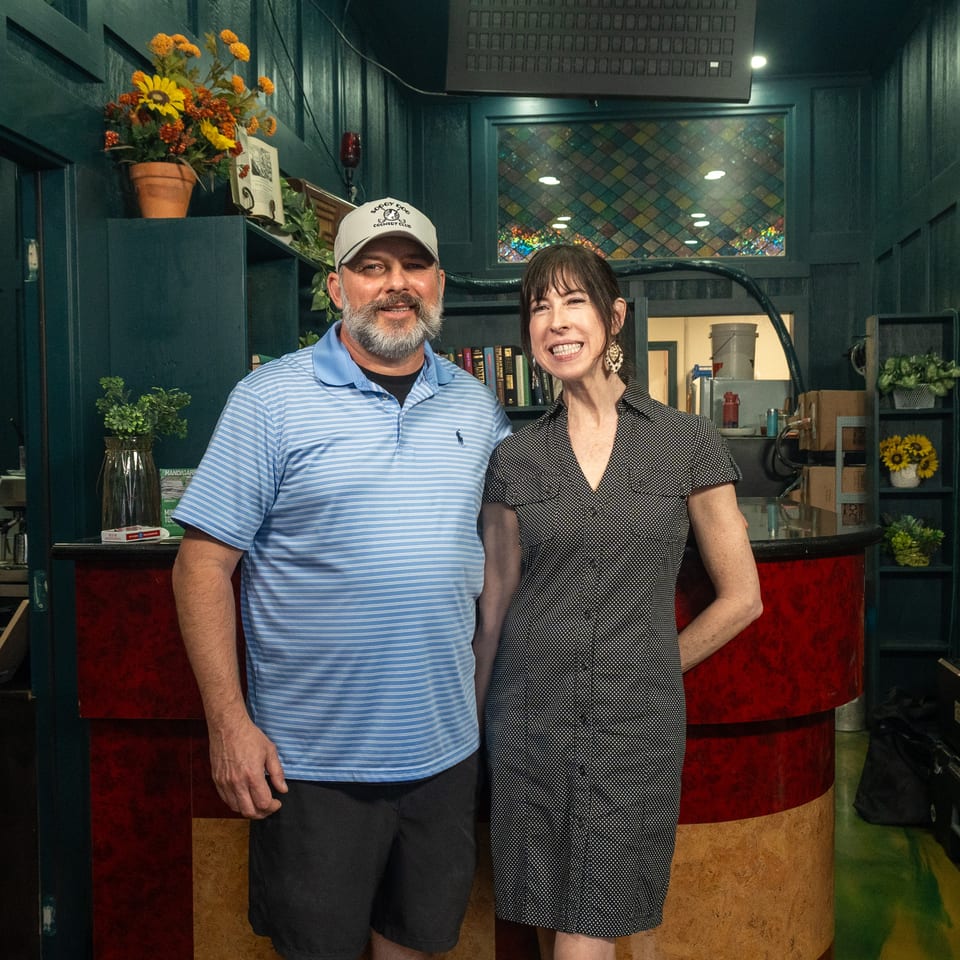 Two smiling people pose together behind a restaurant bar decorated with flowers, books, stained glass, and dark teal walls.