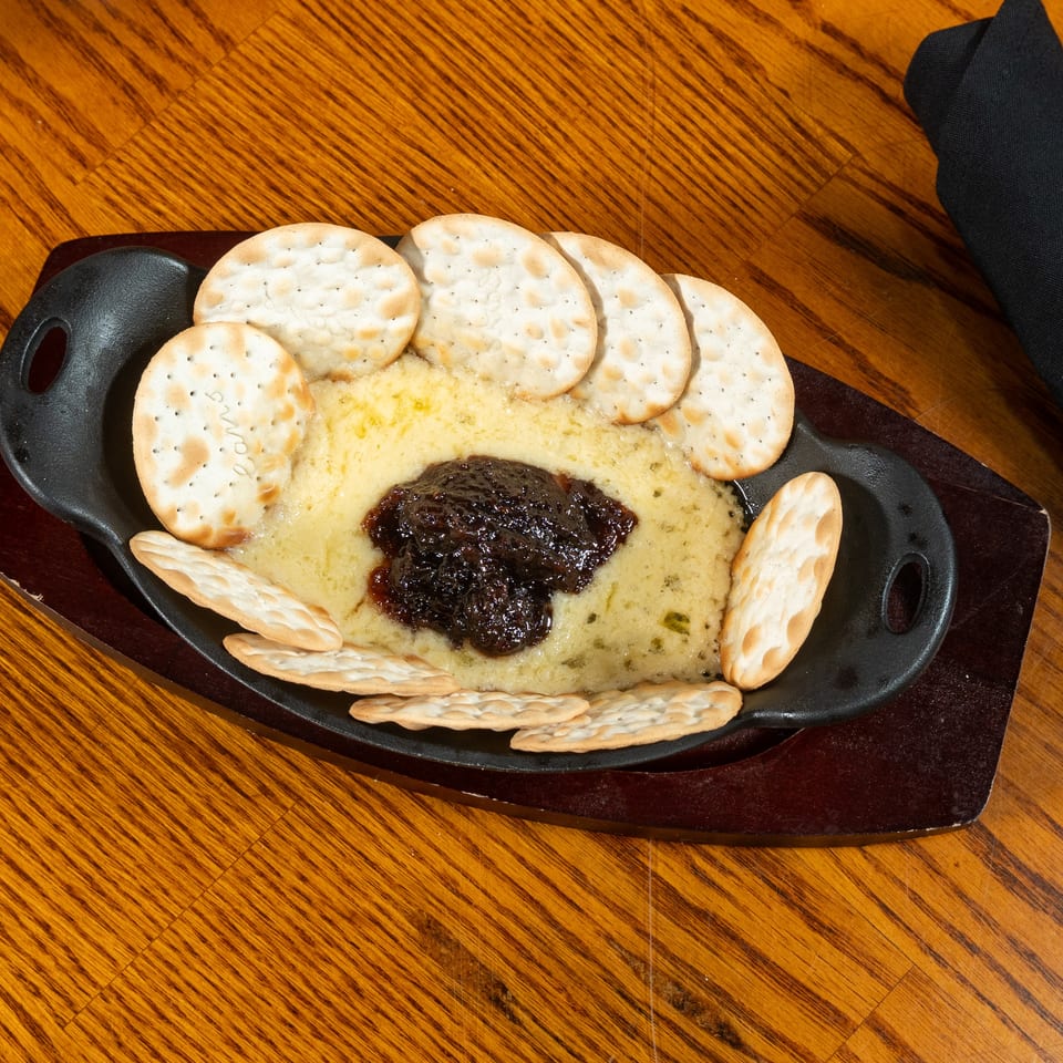 Skillet appetizer with melted cheese, dark dipping sauce, and assorted crackers on a wooden restaurant table.