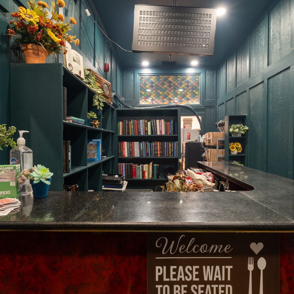 Restaurant host stand with a black countertop, teal walls, bookshelves, flowers, stained-glass window, and welcome sign.