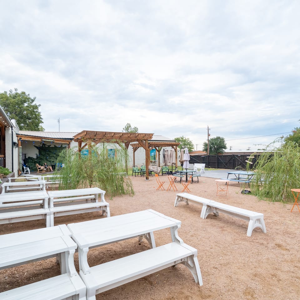 Spacious restaurant patio with white picnic tables, colorful chairs, umbrellas, wooden pergolas, string lights, and leafy landscaping.