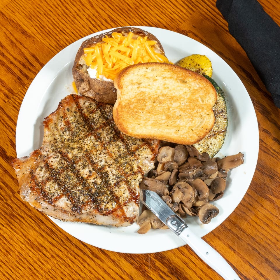 Grilled seasoned steak served with sautéed mushrooms, a cheese-topped baked potato, toasted bread, and vegetables on a white plate.