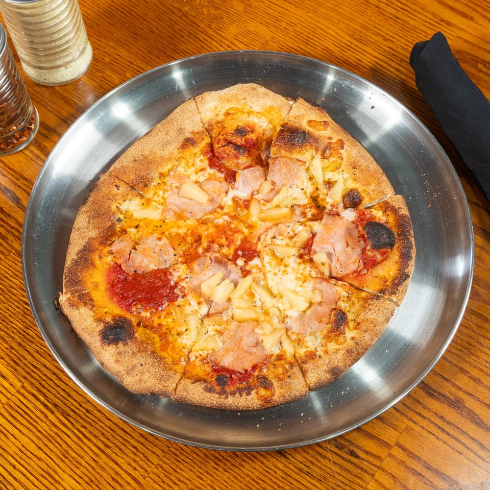 Cheesy ham and pineapple pizza with a charred crust served on a metal tray atop a wooden restaurant table.