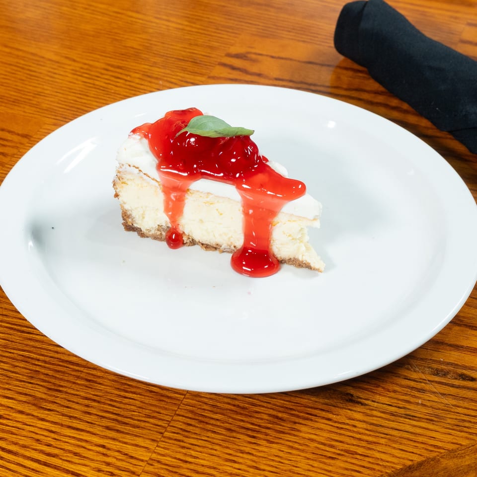 Slice of cheesecake topped with glossy strawberry sauce and a basil leaf on a white plate beside a black napkin.
