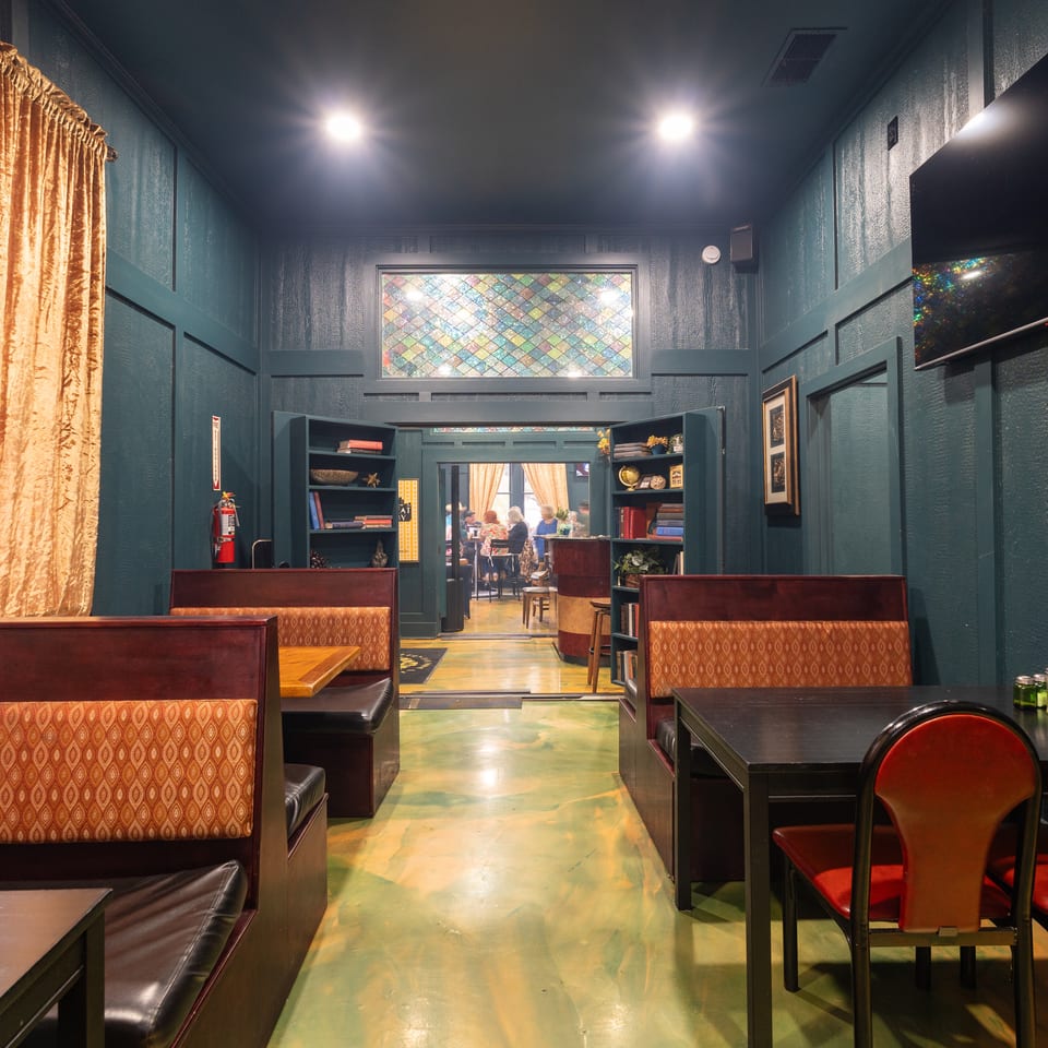 Elegant restaurant dining room with teal walls, patterned booths, red chairs, bookshelves, stained-glass windows, and diners visible beyond.