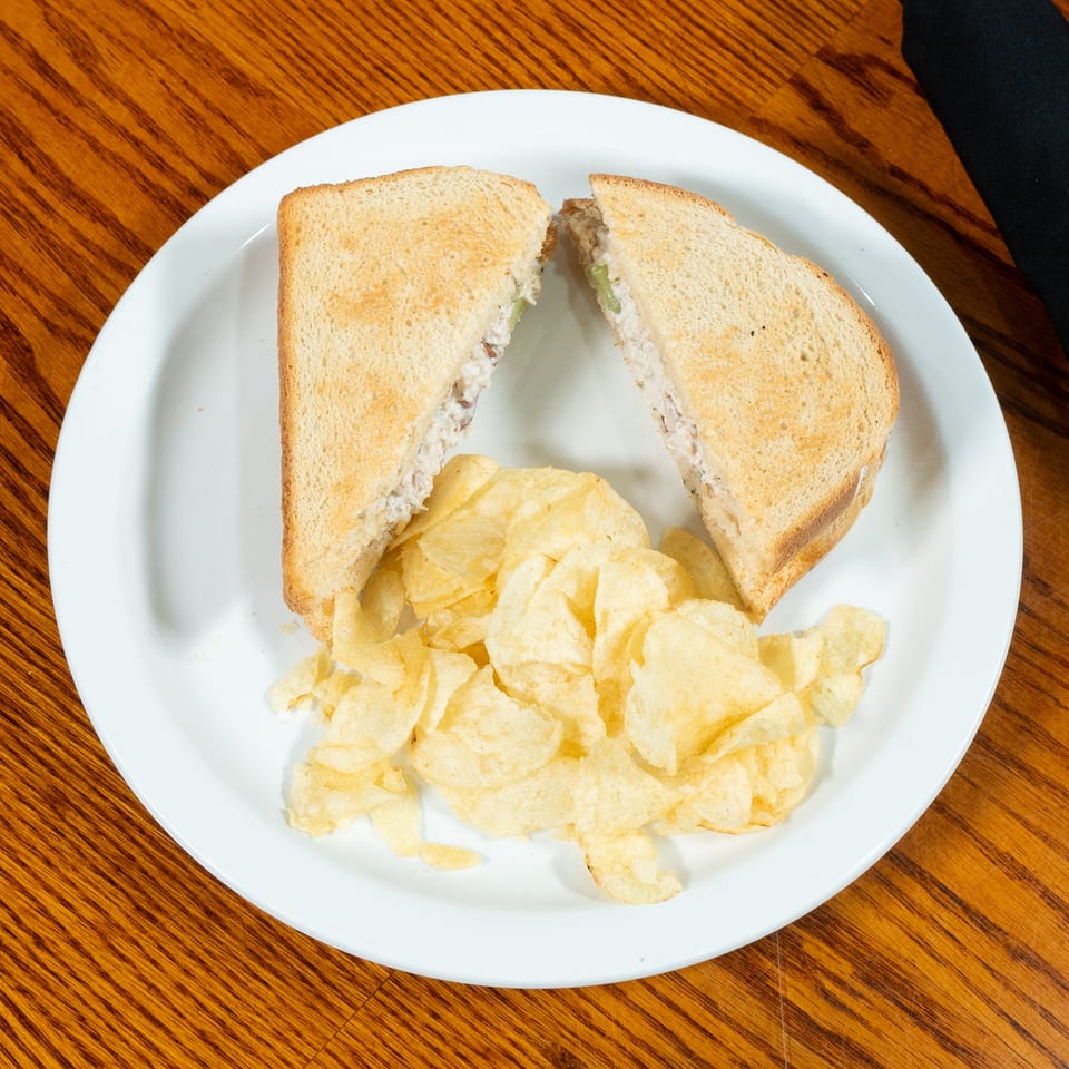 Toasted tuna salad sandwich halves served with potato chips on a white plate atop a wooden table.