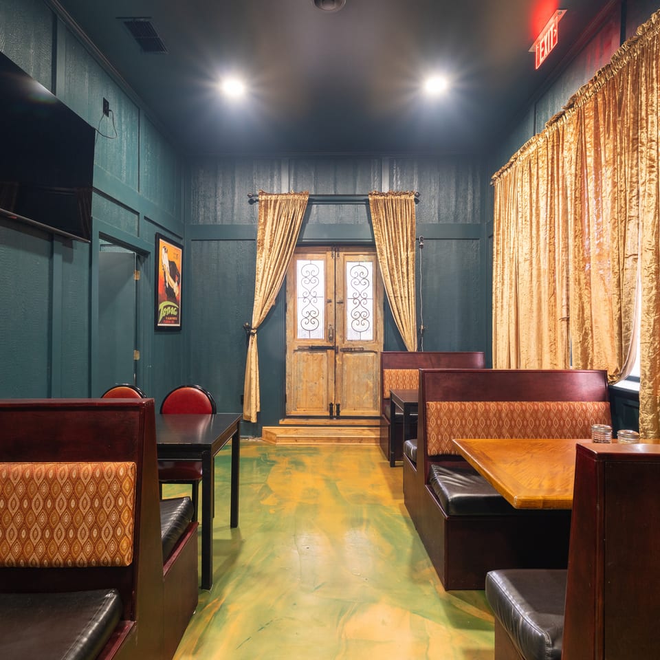 Restaurant dining room with dark teal walls, patterned booths, golden curtains, wooden tables, and a vintage double door.