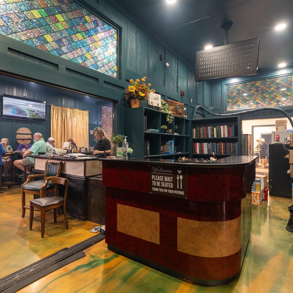 Restaurant host stand with a welcome sign, colorful stained-glass windows, bookshelves, floral decor, and diners seated nearby.