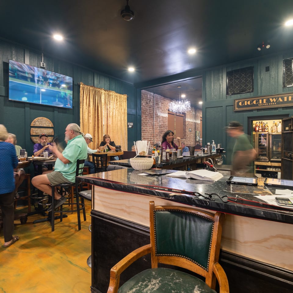 Bustling Gigglewater bar interior with seated patrons, bartending counter, liquor shelves, gold curtains, and television showing sports.