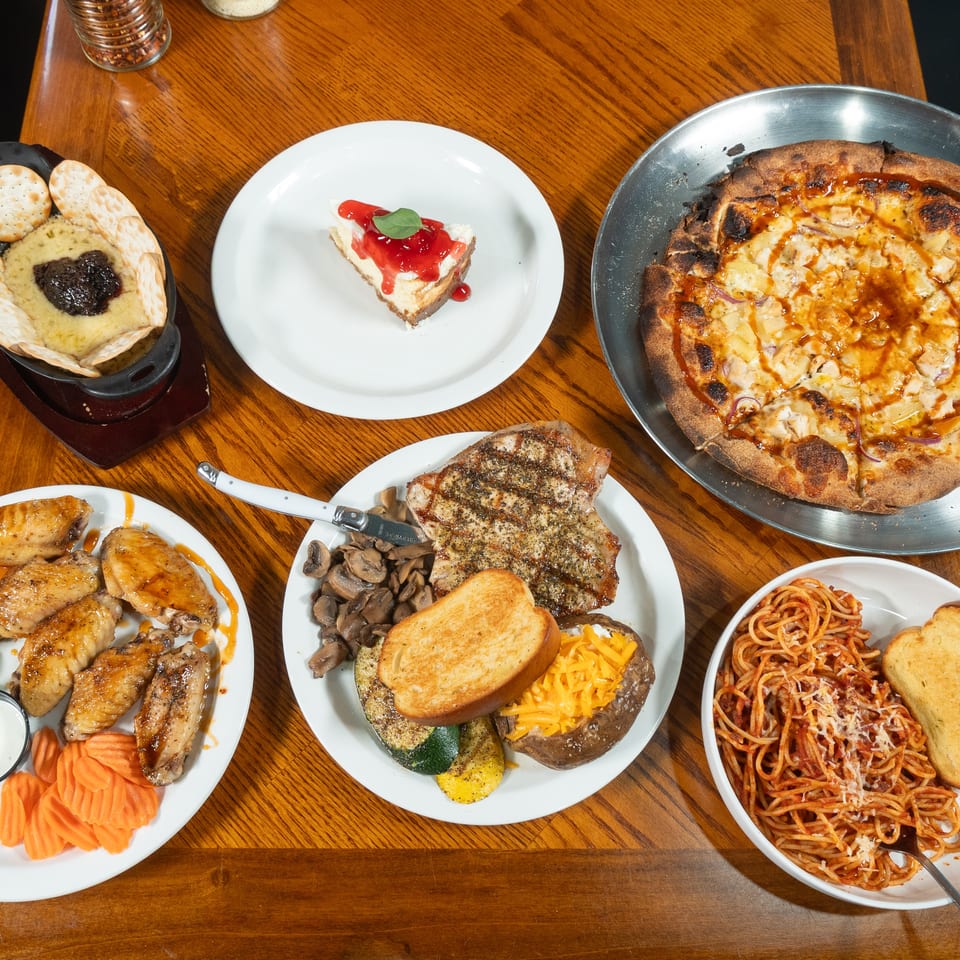 Overhead view of a restaurant table featuring pizza, steak, wings, spaghetti, vegetables, crackers with dip, and cheesecake.
