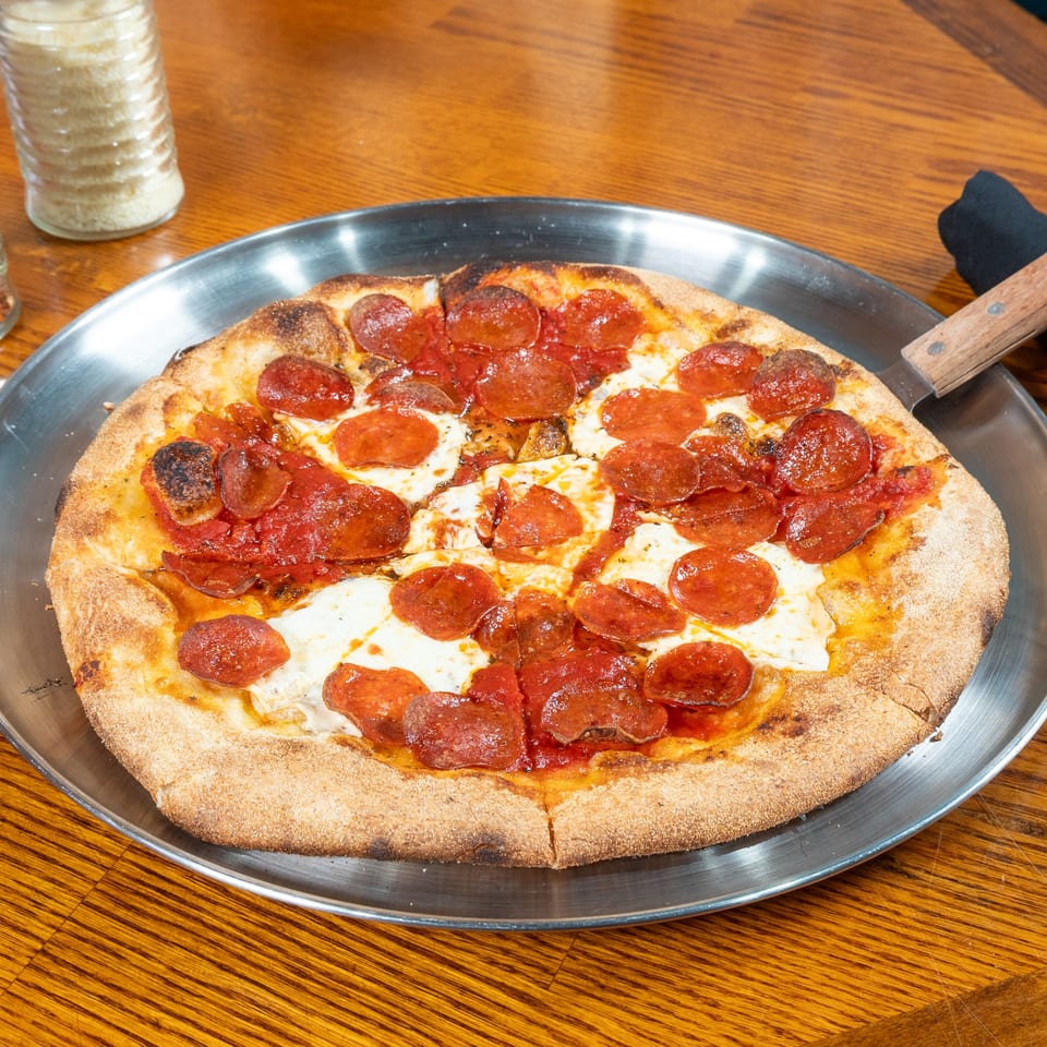 Pepperoni pizza with melted cheese and a crisp crust on a metal tray, accompanied by red pepper flakes and grated cheese.