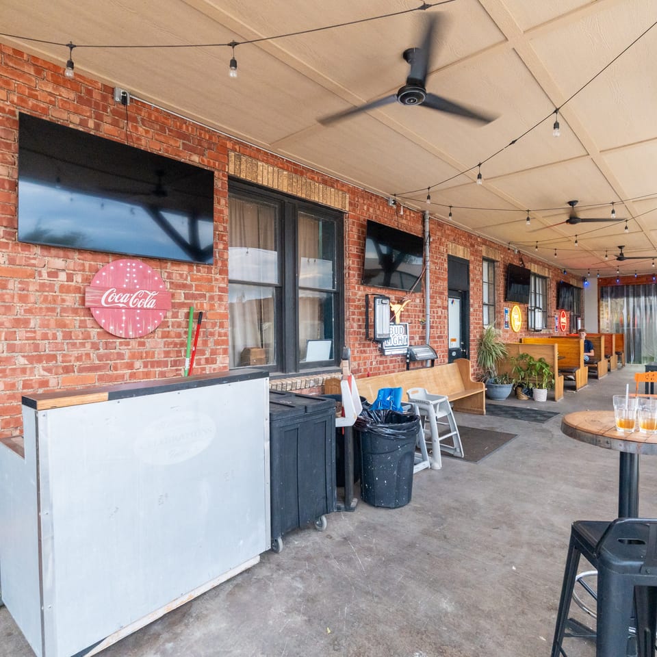 Covered restaurant patio with exposed brick walls, string lights, ceiling fans, colorful signs, booths, tables, and seated diners.