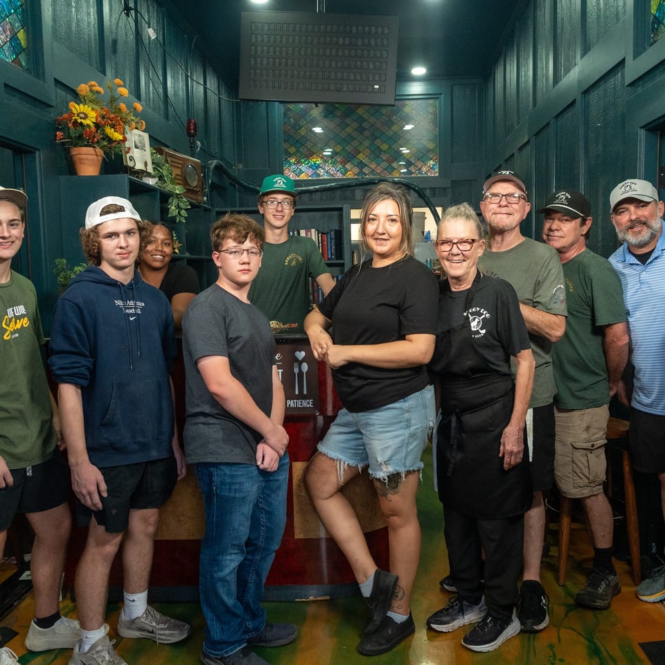 A diverse group poses together inside a warmly decorated restaurant with stained-glass windows, shelves, and colorful flooring.