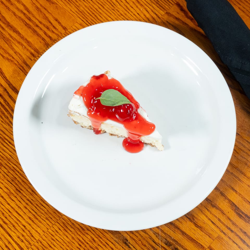 Slice of cheesecake topped with glossy red berry sauce and a green leaf, served on a white plate over a wooden table.